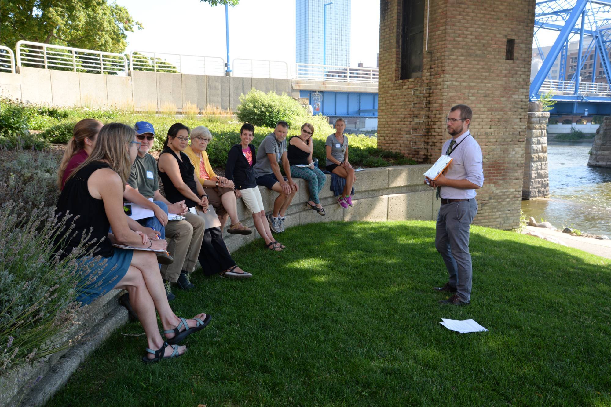 Teachers sitting on a ledge by the grand river listening to a presenter